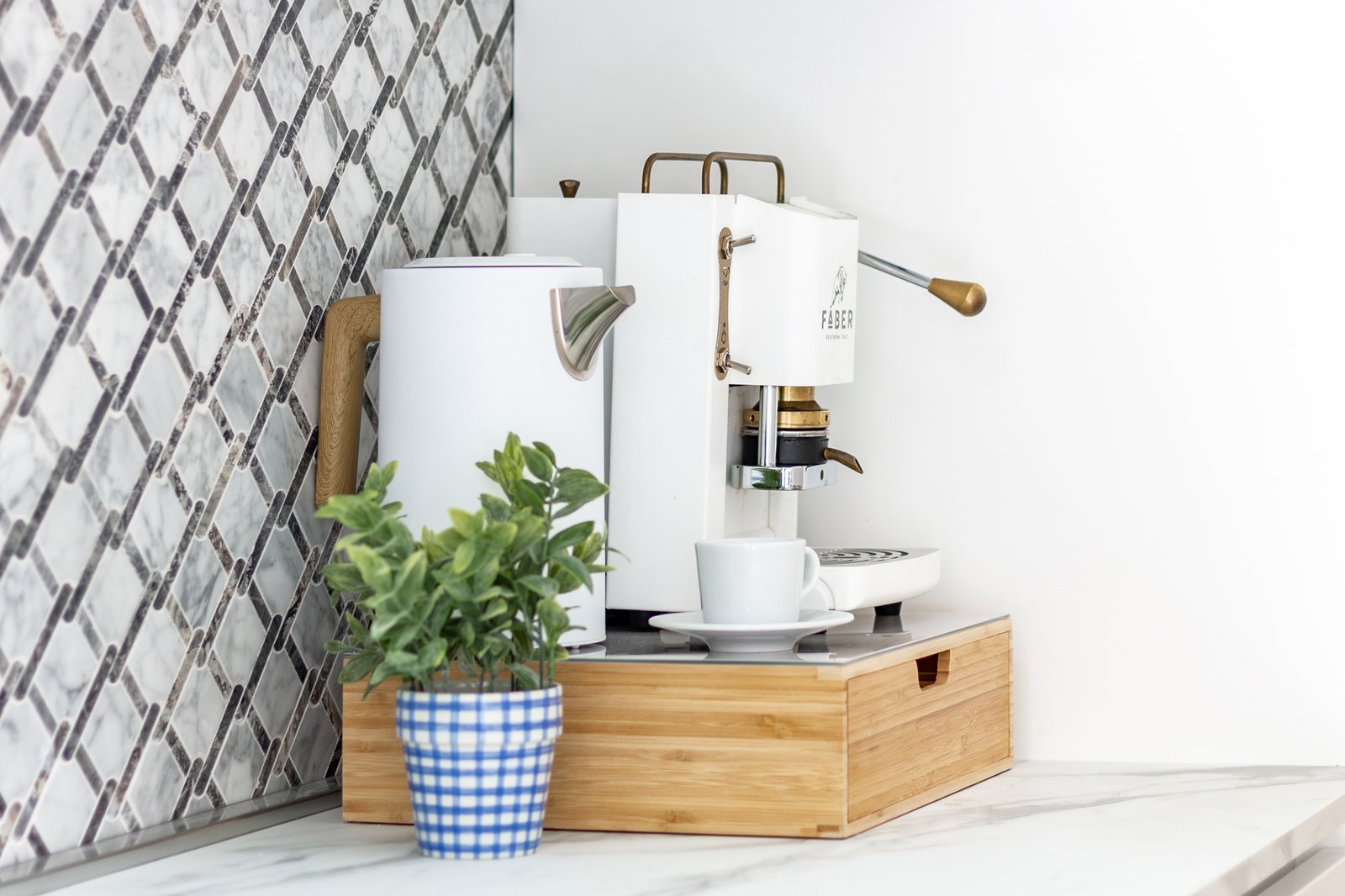 10_angolo-caffe-moderno-con-macchina-bianca-e-piantina.jpg Angolo caffè moderno e luminoso con macchina espresso bianca, bollitore bianco con manico in legno e piantina verde in vaso a quadretti blu, su piano in marmo e sfondo con piastrelle geometriche grigie.