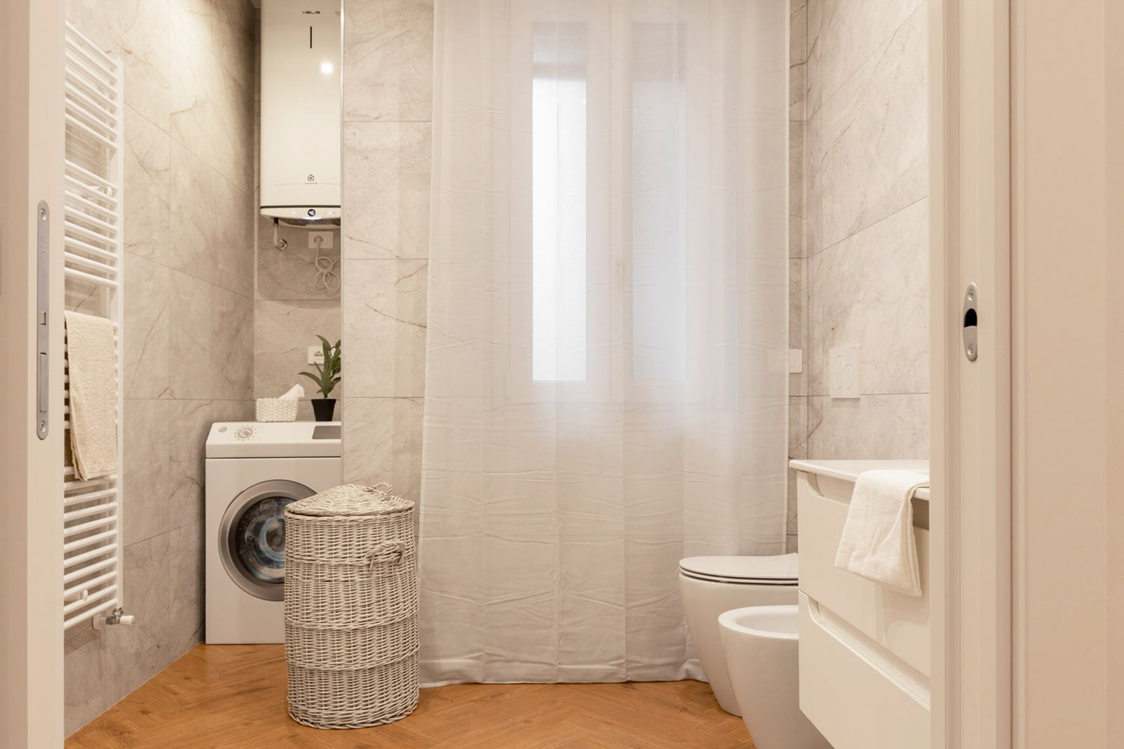 10_home-staging-bagno-moderno-minimalista-marmo-toni-neutri-pavimento-herringbone.jpg Bagno moderno e minimalista con pavimento in legno a spina di pesce e pareti effetto marmo; lavatrice bianca sotto scaldabagno, cesto portabiancheria in vimini e finestra con tende bianche, sanitari e mobile lavabo chiari; ambiente luminoso.