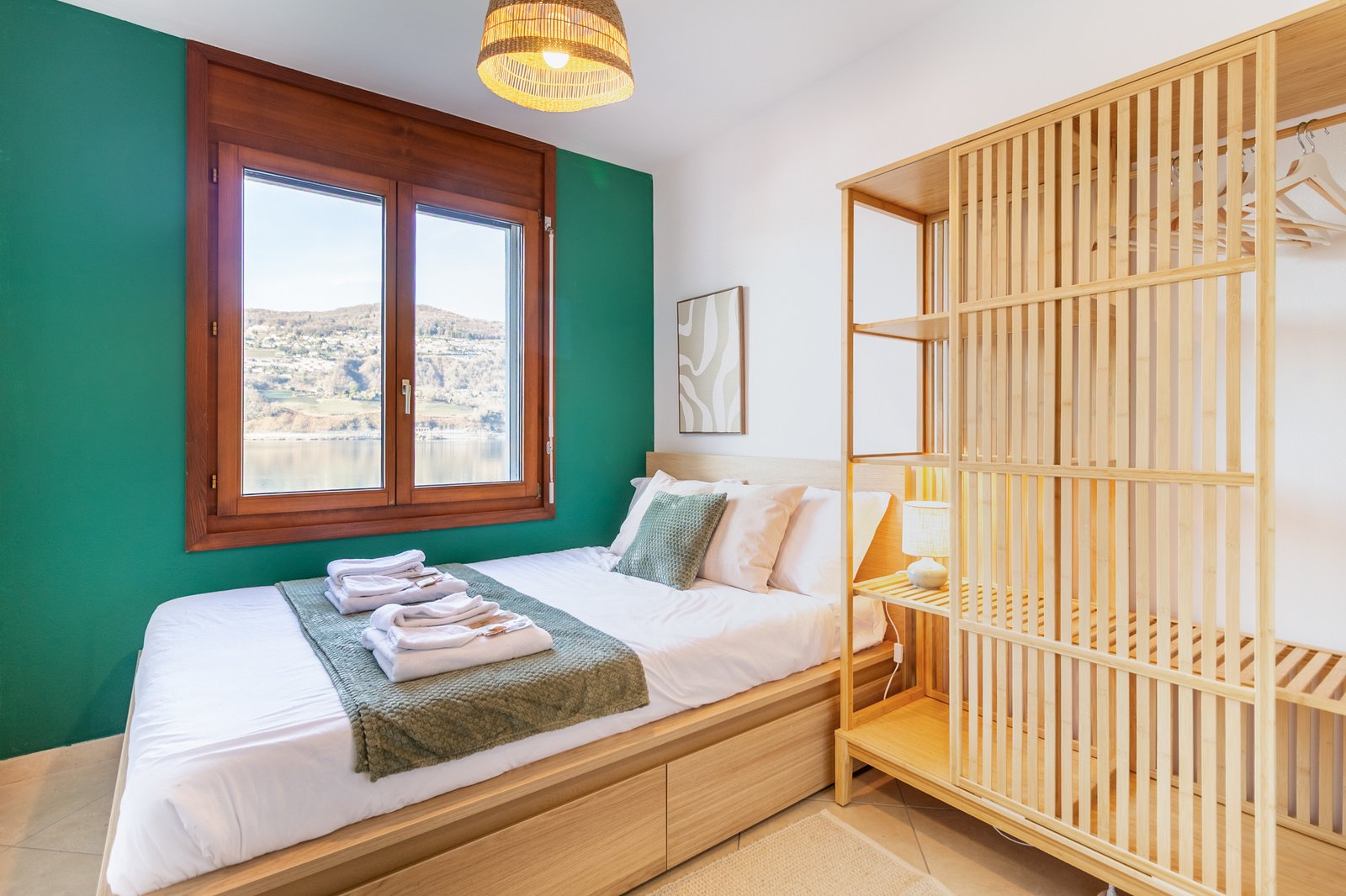 10_relooking-appartamento-camera-matrimoniale-armadio-legno-design-minimal-caldo-parete-verde-vista-lago.jpg Camera con armadio aperto in legno, parete verde e finestra panoramica sul lago, luce naturale e comfort; complementi verdi e beige; toni neutri e atmosfera rilassata.