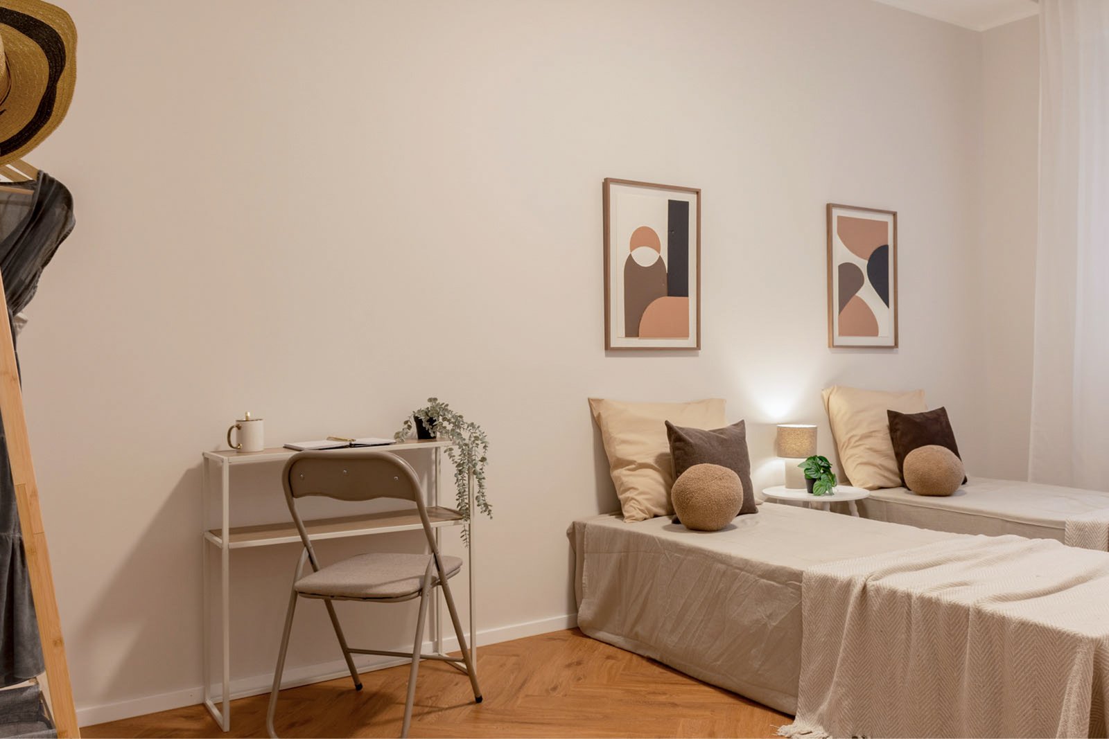 11_home-staging-camera-doppia-moderno-minimalista-toni-neutri-pavimento-herringbone.jpg Camera doppia moderna e minimalista con due letti singoli rivestiti in tessuti beige e cuscini marroni; tra i letti comodino con lampada e pianta verde, parete con quadri astratti dai toni caldi, angolo studio con scrivania bianca e sedia pieghevole, pavimento in legno a spina di pesce e decorazioni naturali.