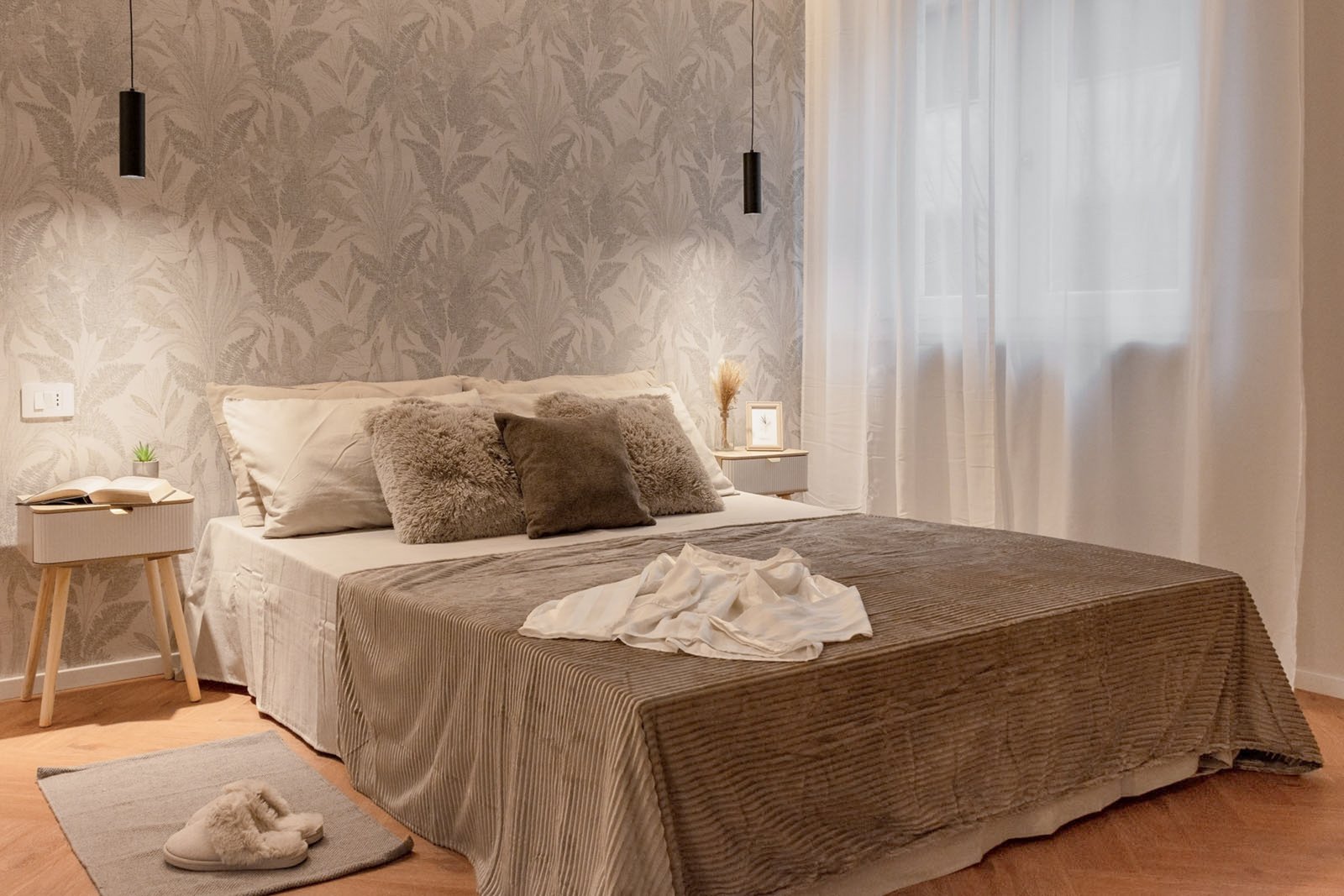 13_home-staging-camera-matrimoniale-moderno-minimalista-toni-neutri-pavimento-herringbone.jpg Camera matrimoniale moderna e minimalista con parete decorata da carta da parati botanica tortora, letto con copriletto beige e cuscini in pelliccia e tessuti neutri; comodini in legno chiaro con lampade sospese nere, tappeto e pantofole sul pavimento in legno a spina di pesce, finestra con tende bianche che diffondono luce naturale, ambiente elegante.