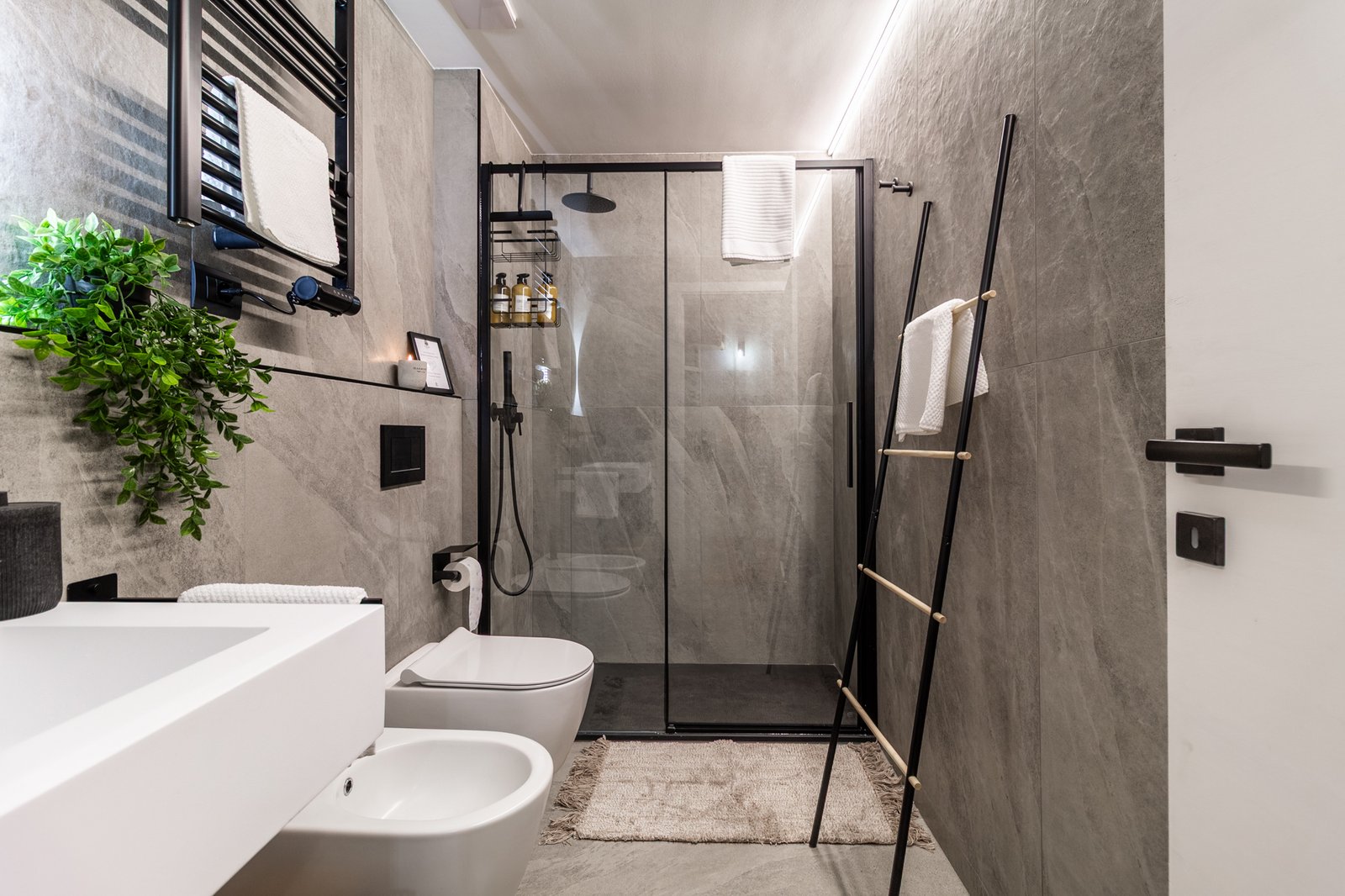 13_relooking-appartamento-montagna-bagno-moderno-effetto-pietra-grigio-nero-design-minimal-elegante.jpg Bagno moderno con pareti effetto pietra grigia, doccia con vetro e finiture nere, e sanitari bianchi, atmosfera elegante e minimalista con dettagli curati e luce soffusa.