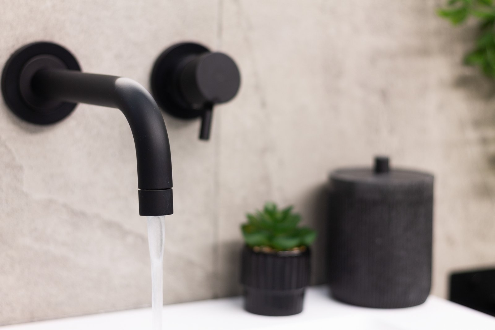 14_dettaglio-bagno-rubinetto-nero-design-moderno-minimal.jpg Dettaglio di rubinetto nero opaco con acqua corrente su lavabo bianco, sfondo effetto pietra e decorazioni minimal; design moderno e dettagli curati.