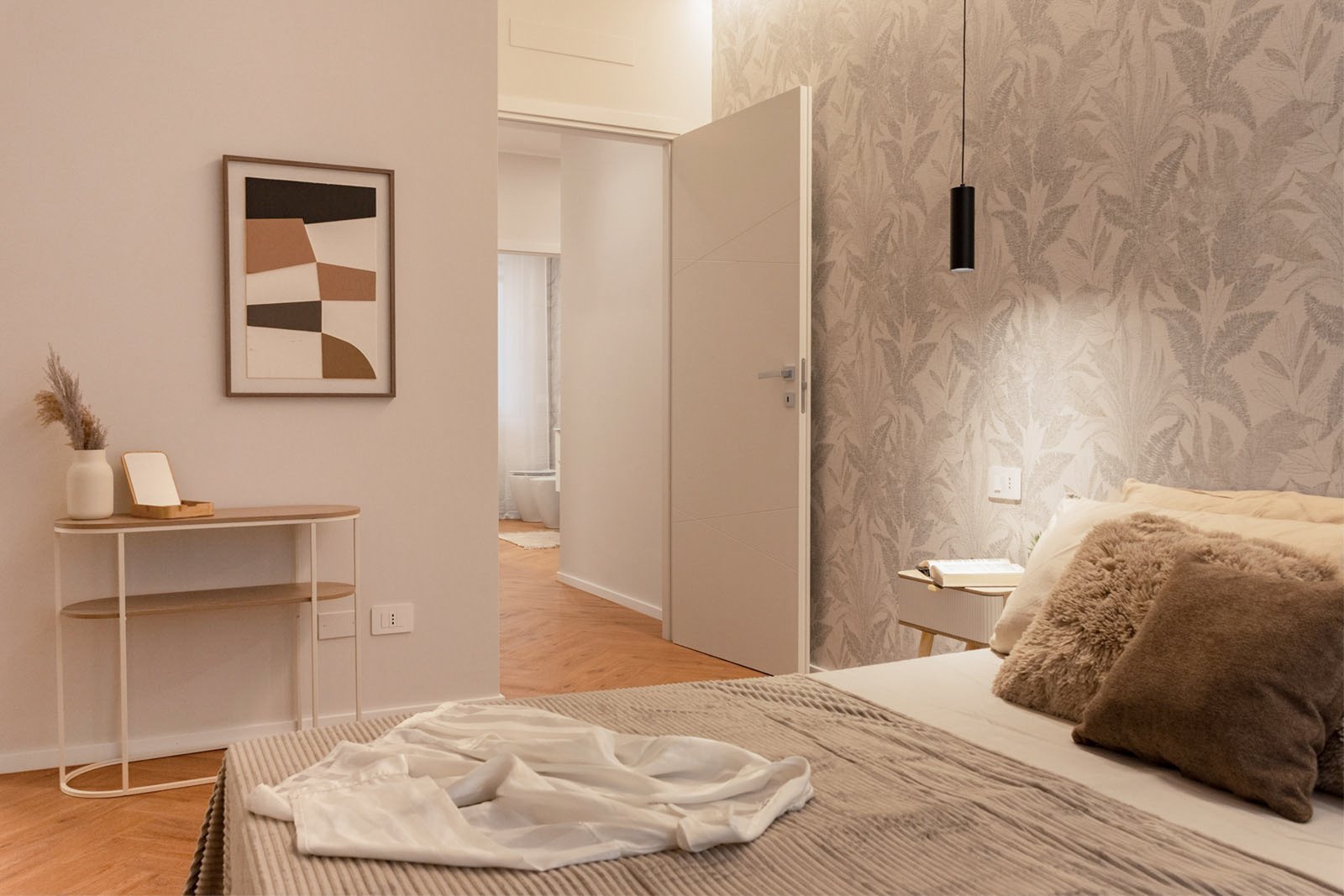 14_home-staging-camera-matrimoniale-moderno-minimalista-toni-neutri-legno-pavimento-herringbone.jpg Camera matrimoniale moderna e minimalista con parete decorata da carta da parati botanica tortora, letto con copriletto beige e cuscini in pelliccia e tessuti neutri; comodino in legno chiaro con lampada sospesa nera, quadro astratto dai toni caldi e consolle con decorazioni naturali, pavimento in legno a spina di pesce; illuminazione morbida e ambiente elegante.