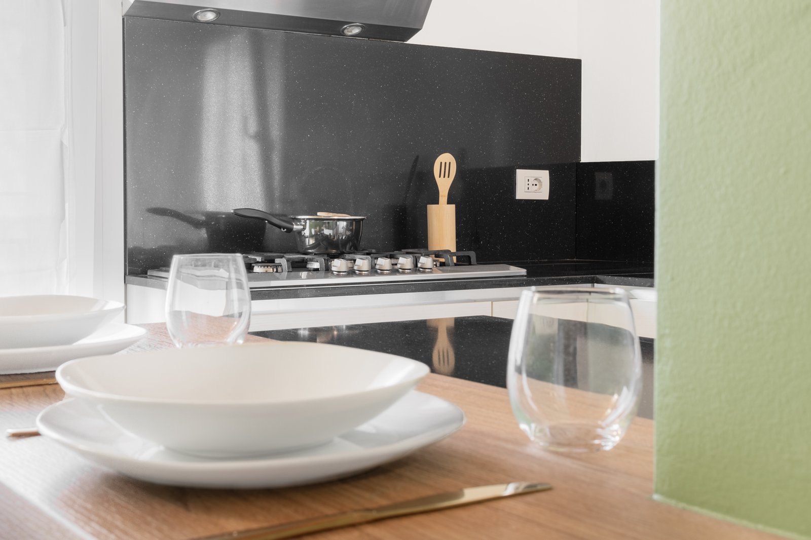 14_home-staging-cucina-mansarda-dettaglio-isola-apparecchiata-fornelli-neri.jpg Dettaglio della cucina mansardata con colonna color salvia e isola apparecchiata con piatti bianchi moderni e bicchieri in vetro. Sullo sfondo, il blocco cottura con pannelli neri è a fuoco, con pentola sui fornelli e portamestoli in legno, in un insieme elegante e contemporaneo.