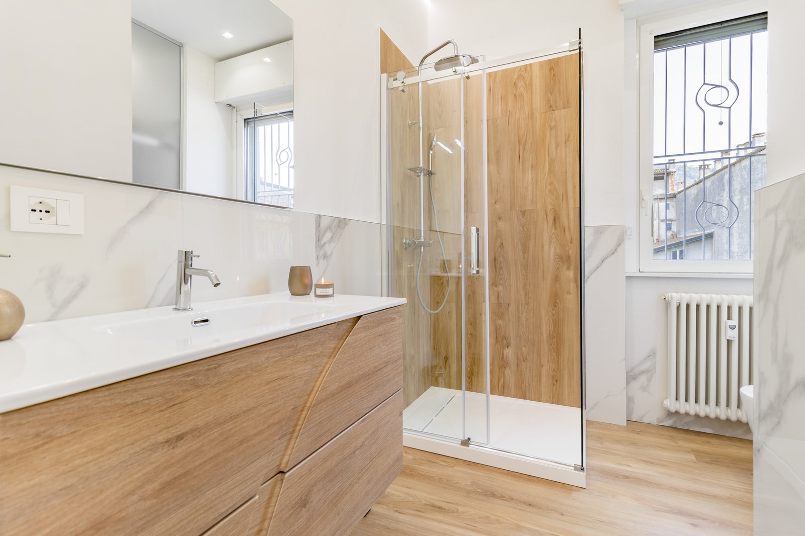 15_bagno-moderno-con-mobili-in-legno-e-box-doccia-vetro.jpg Bagno moderno e luminoso con mobile in legno chiaro, lavabo bianco con rubinetto cromato, box doccia in vetro con pareti effetto legno e pavimento coordinato.