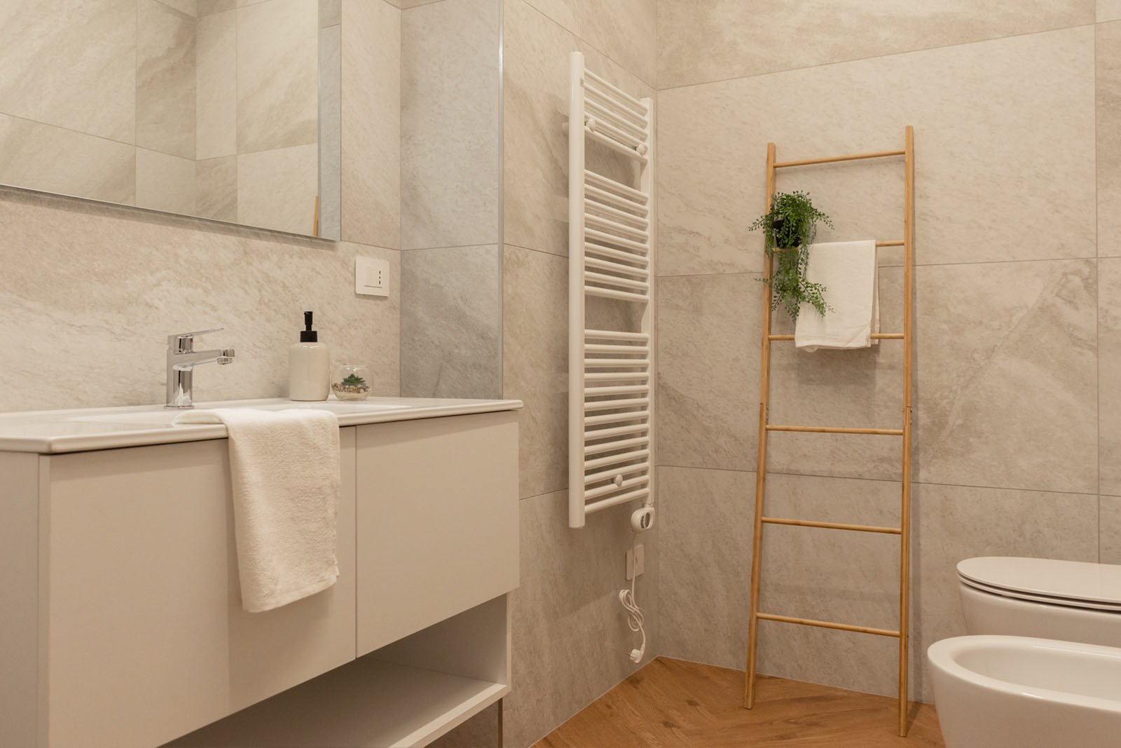 15_home-staging-bagno-moderno-minimalista-marmo-legno-pavimento-herringbone.jpg Bagno moderno e minimalista con pareti rivestite in piastrelle effetto marmo grigio e pavimento in legno a spina di pesce; mobile lavabo sospeso bianco con asciugamano e dispenser, specchio ampio, scaldasalviette bianco e scala in legno con pianta verde e asciugamano.