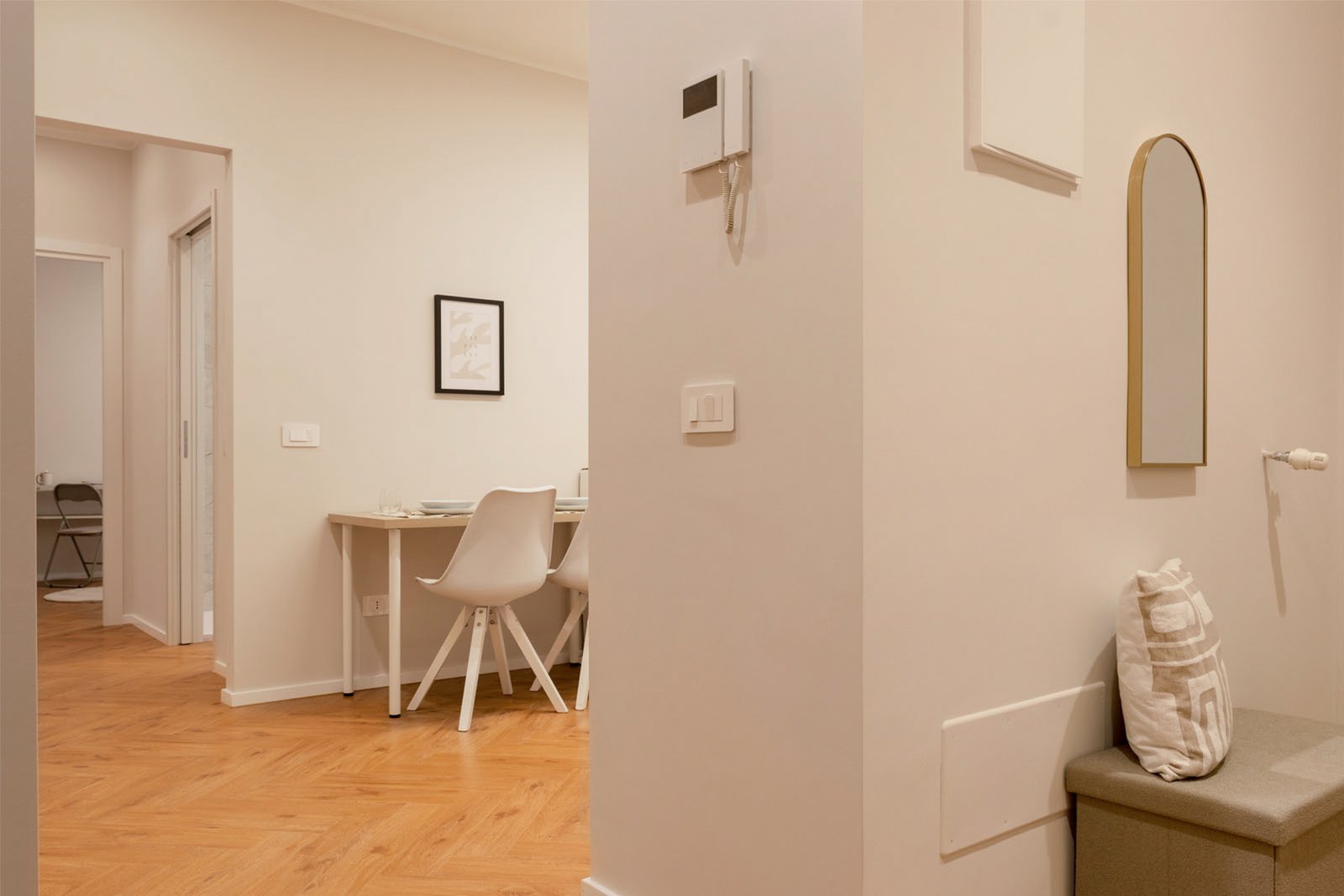 1_home-staging-ingresso-moderno-minimalista-pavimento-legno-herringbone.jpg Interno moderno e minimalista con pavimento in legno a spina di pesce, pareti chiare e arredi essenziali; in primo piano una panca con cuscino geometrico e specchio ad arco dorato, sullo sfondo tavolo da pranzo con sedie bianche e quadro astratto.