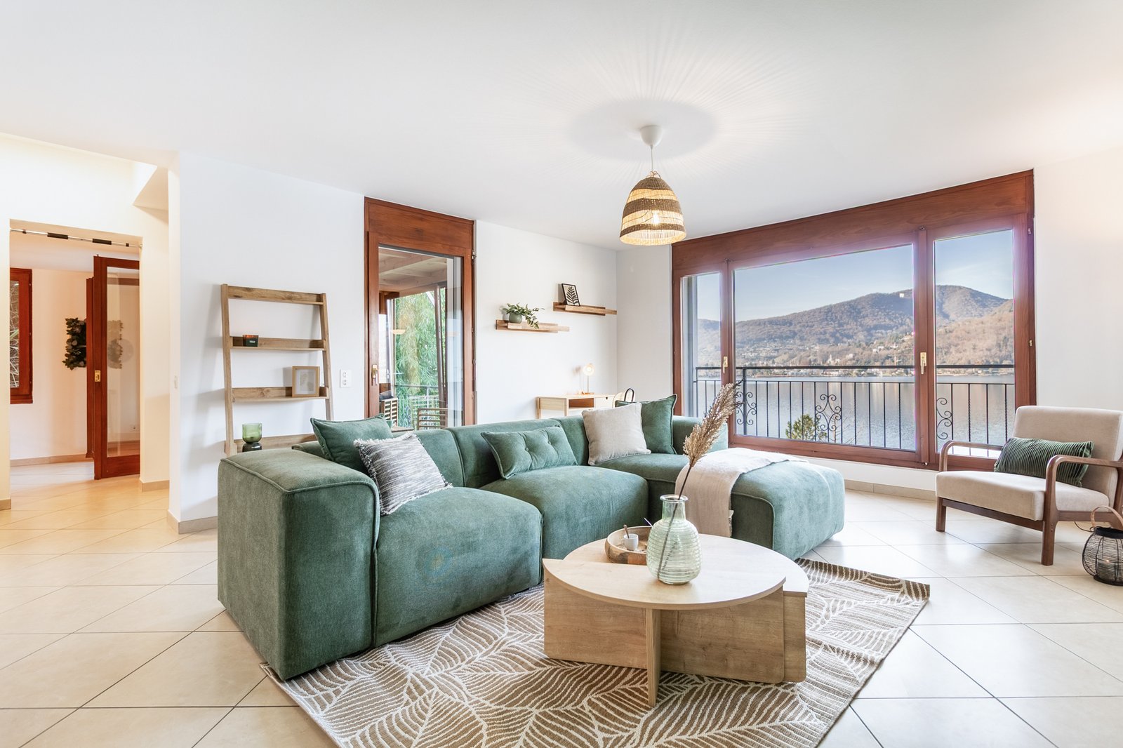 Soggiorno moderno con divano verde, poltrona beige e grandi vetrate sul lago, luce naturale e design elegante.
