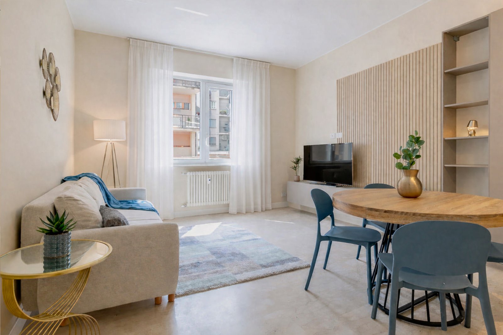 Soggiorno moderno e luminoso con arredamento minimalista, pareti chiare, tavolo rotondo in legno, sedie blu e divano beige con plaid azzurro.