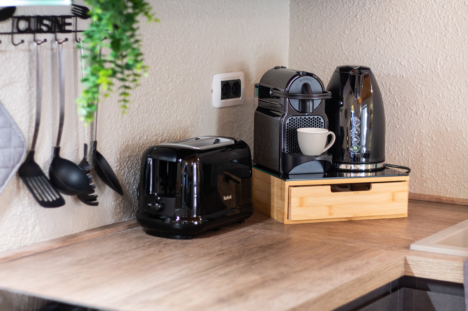 2_dettaglio-cucina-moderno-relooking-appartamento-design.jpg Angolo cucina moderno con tostapane, macchina da caffè e bollitore neri su piano in legno; appartamento curato e dal design elegante.