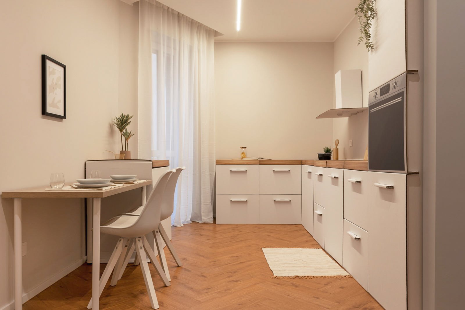 2_home-staging-cucina-moderna-minimalista-legno-bianco-pavimento-legno-herringbone.jpg Cucina moderna e minimalista con pavimento a spina di pesce; tavolo da pranzo con due sedie bianche, piatti apparecchiati. Luce LED e naturale filtrata da tende bianche.