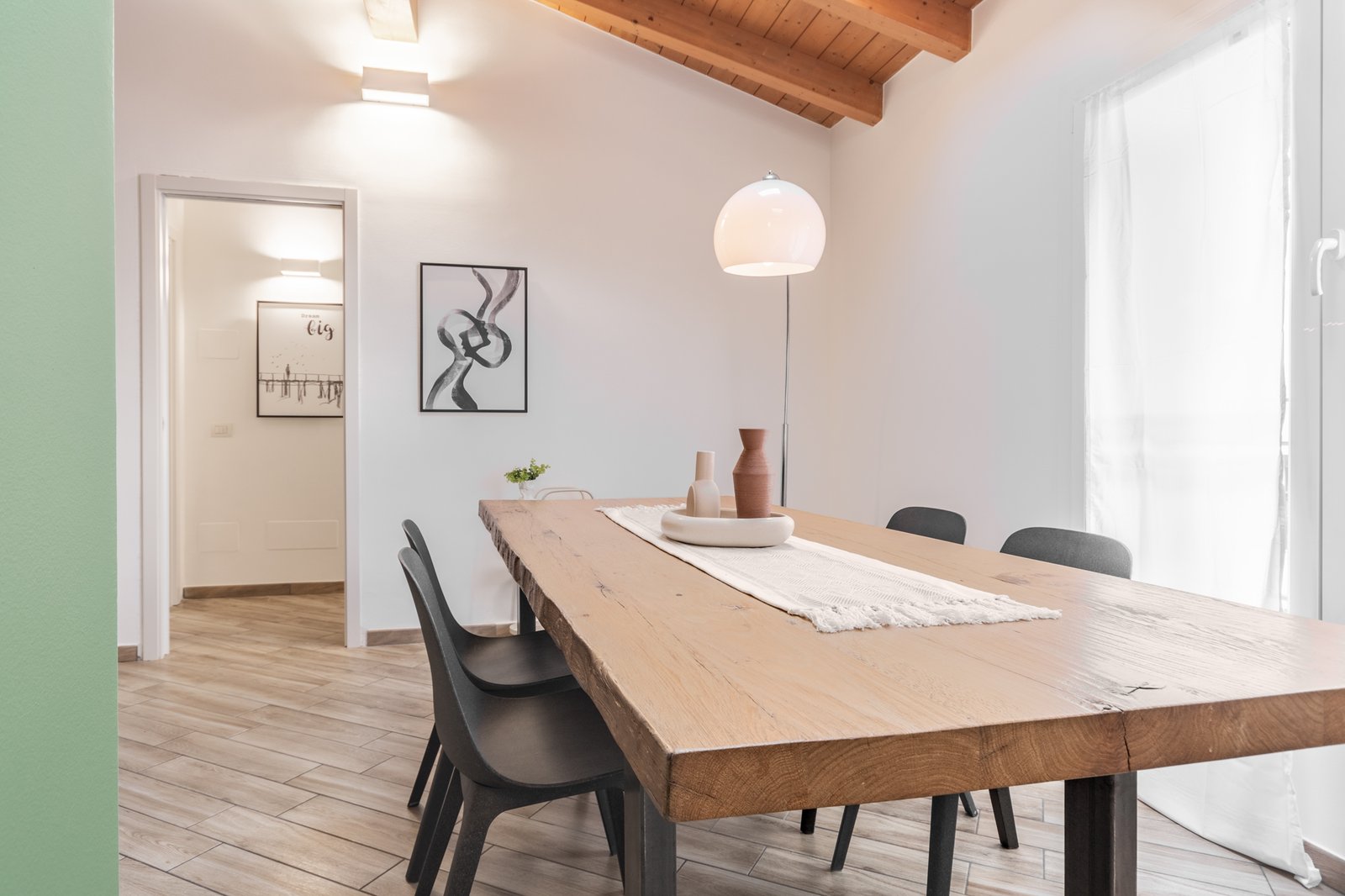 2_home-staging-mansarda-zona-pranzo-tavolo-legno-ferro-lampada-arco.jpg Dettaglio della zona pranzo in una mansarda luminosa, con tavolo in legno e gambe in ferro battuto, sedie nere e runner beige con centrotavola minimalista, illuminato da una raffinata lampada ad arco bianca e argento e completato da quadro contemporaneo essenziale.