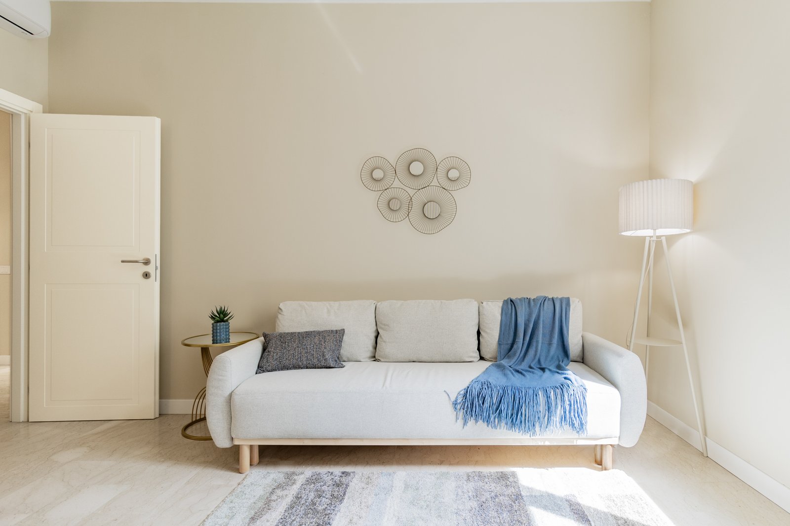 2_relooking-divano-moderno-minimal-parete-beige-cuscini-blu.jpg Divano moderno color beige con cuscino blu scuro e plaid blu, lampada da terra bianca e tavolino dorato con pianta, in soggiorno luminoso dallo stile minimalista e raffinato.