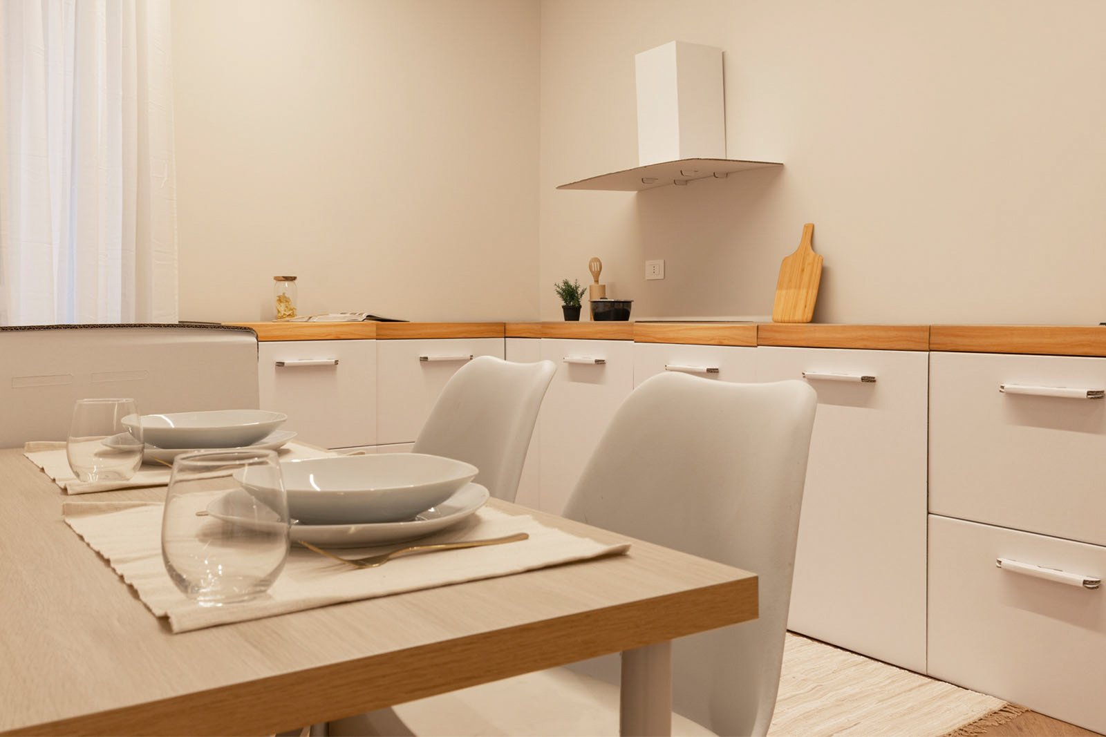 3_home-staging-dettaglio-cucina-moderno-minimalista-legno-bianco-tavolo-apparecchiato.jpg Dettaglio di cucina moderna e minimalista con tavolo apparecchiato per due persone, piatti bianchi e bicchieri in vetro; sullo sfondo cucina in cartone utilizzata per home staging, arricchita da piccoli accessori da cucina.