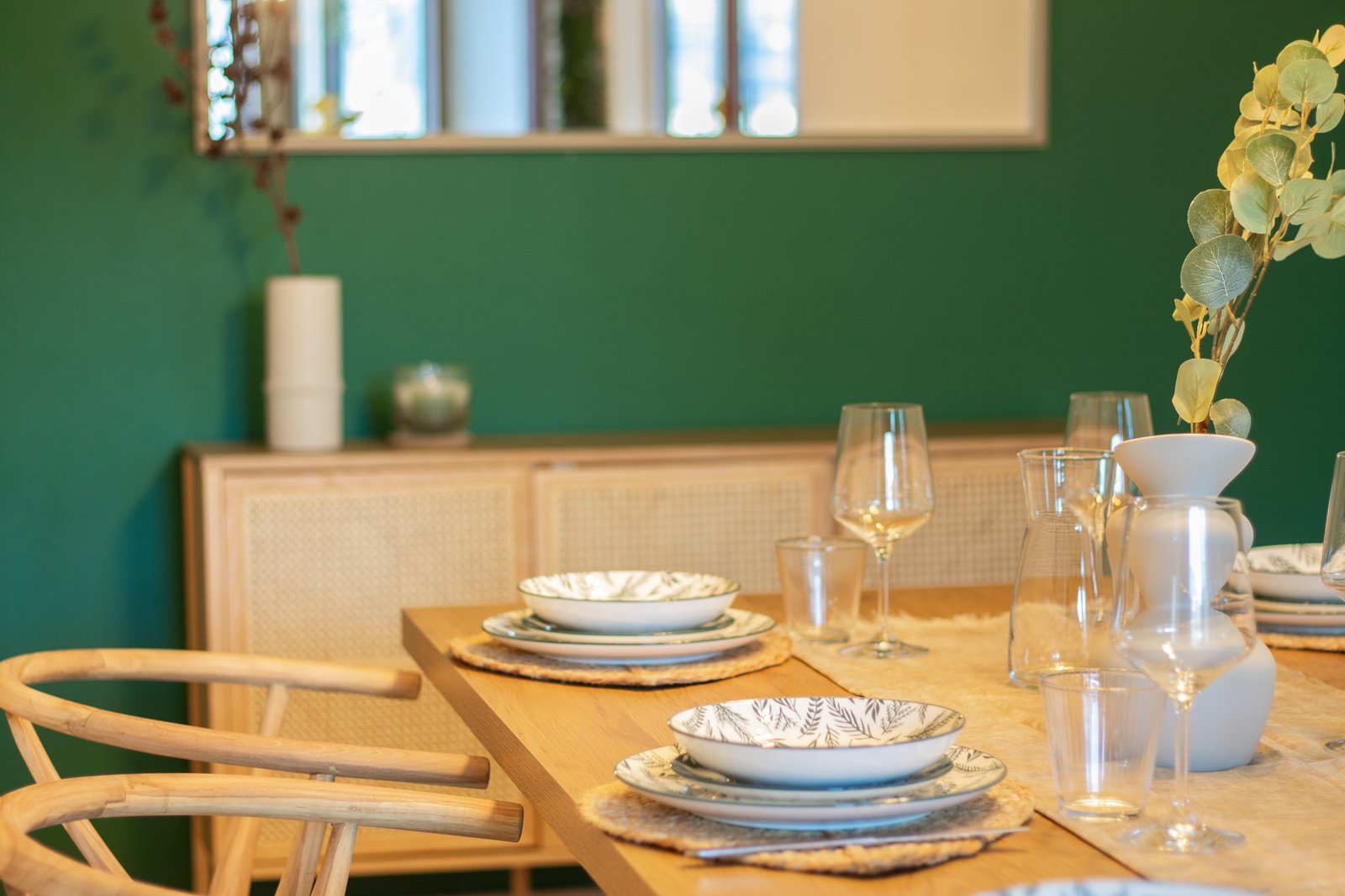 4_dettaglio-zona-pranzo-parete-verde-legno-stile-naturale.jpg Tavolo da pranzo in legno chiaro apparecchiato con piatti decorativi e parete verde, stile naturale e accogliente.