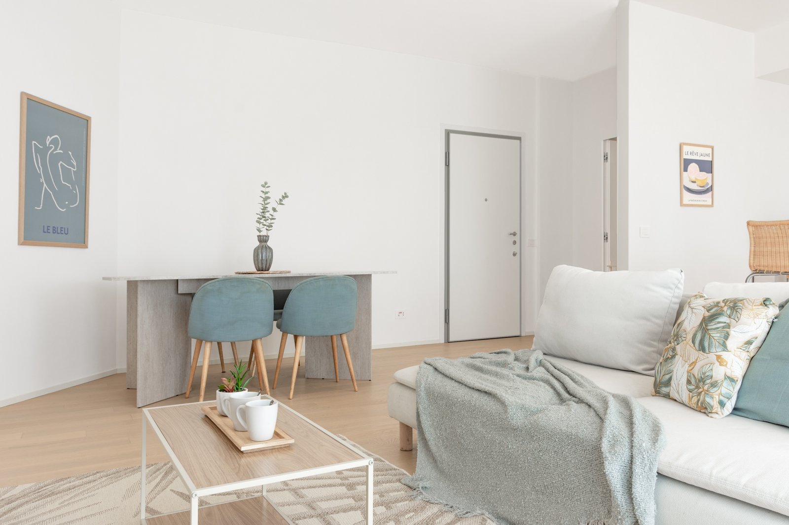 Soggiorno open space moderno e luminoso con divano chiaro, cuscini decorativi, tavolino con struttura bianca in metallo e piano in legno.Zona pranzo con tavolo effetto travertino e sedie azzurre.
