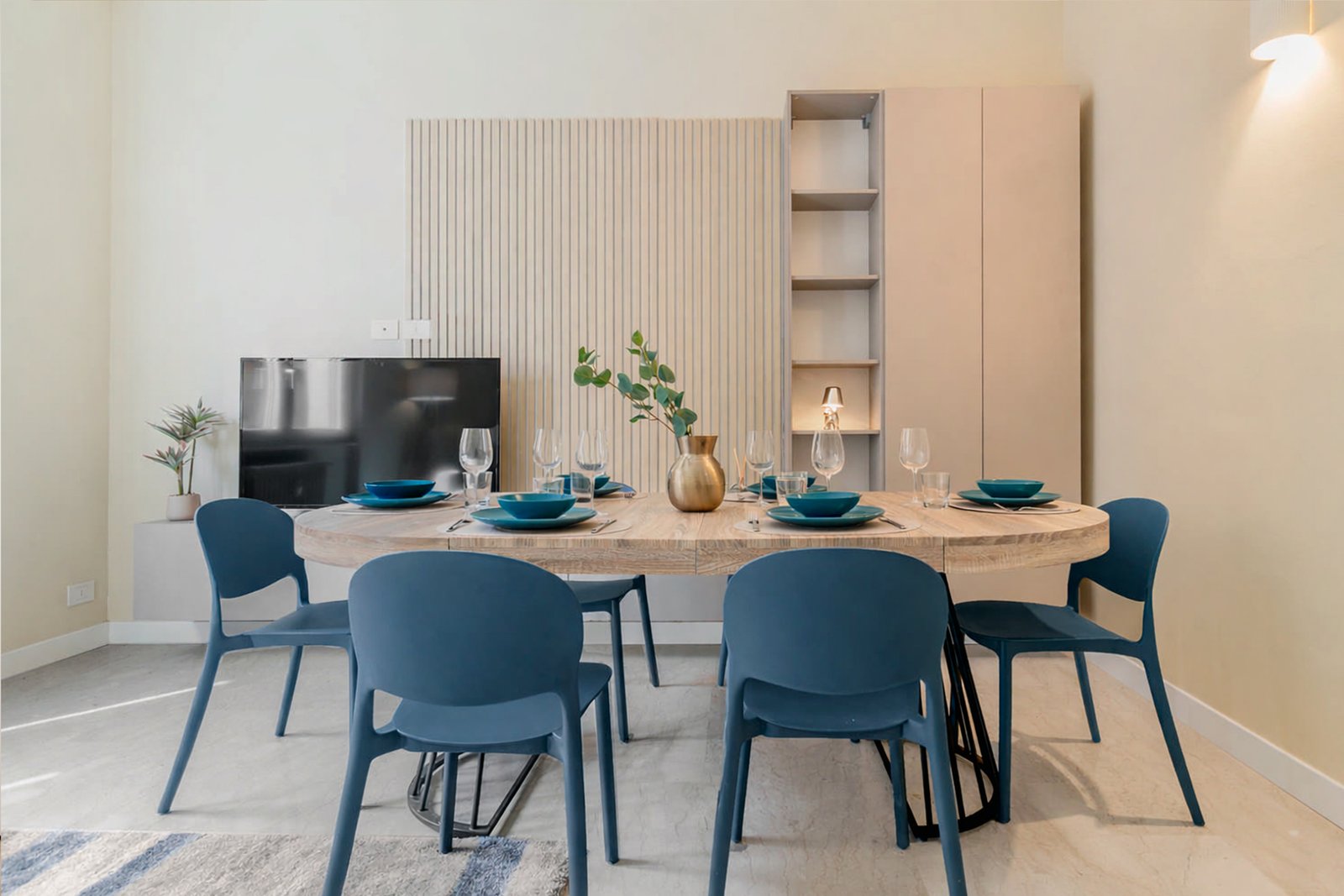 4_relooking-sala-pranzo-moderno-tavolo-in-legno-e-sedie-blu.jpg Sala da pranzo moderna con tavolo rotondo in legno chiaro, sedie blu, mise en place elegante e vaso dorato con pianta verde, in ambiente luminoso e minimalista.