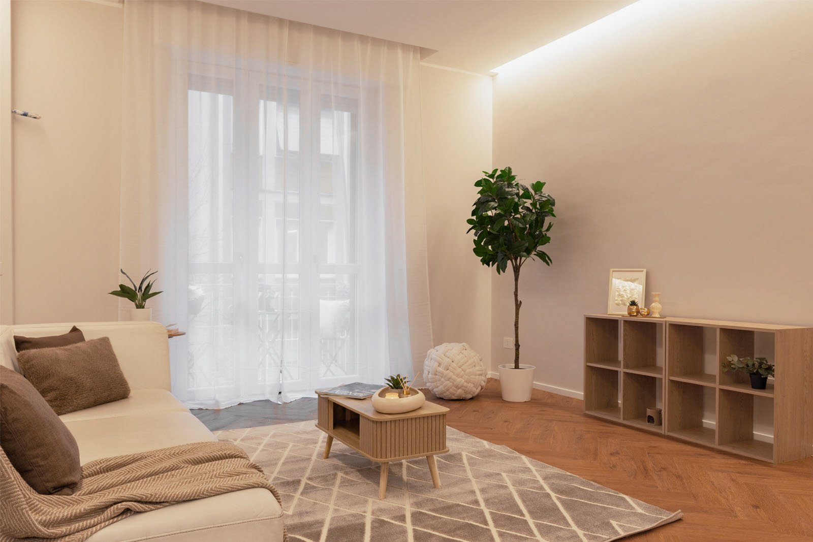 Soggiorno moderno e minimalista con divano bianco e cuscini beige, tavolino in legno chiaro su tappeto geometrico, scaffale con decorazioni e pianta verde accanto alla finestra con tende bianche trasparenti; illuminazione soffusa a LED sul soffitto.