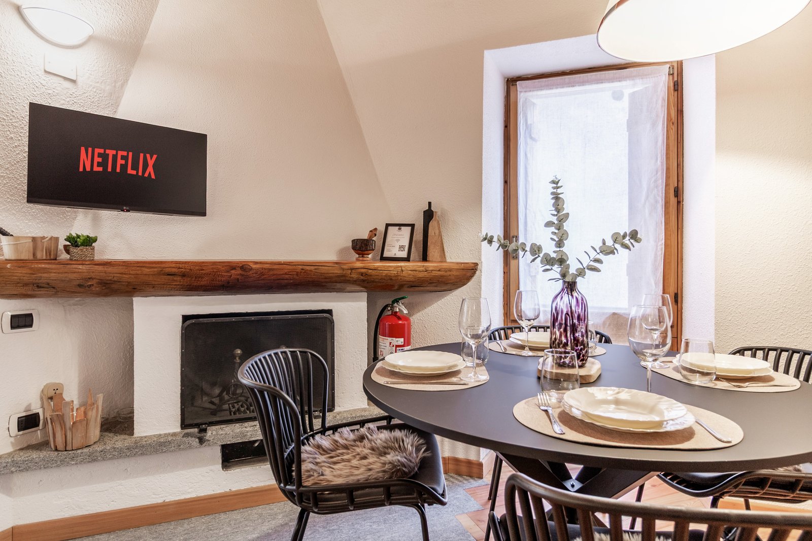 5_relooking-appartamento-moderno-montagna-zona-pranzo-con-camino-design-accogliente.jpg Zona pranzo moderna con camino e tavolo rotondo apparecchiato, ambiente luminoso e accogliente con dettagli in legno e decorazioni elegante.