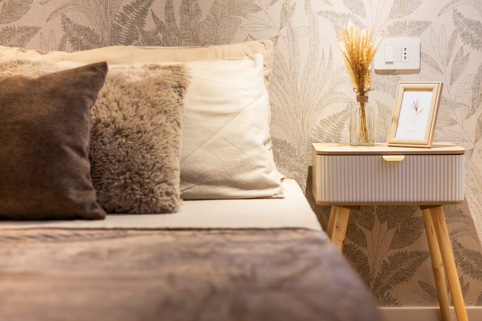 Camera matrimoniale accogliente con tessili beige e marroni, comodino in legno e carta da parati decorativa con motivo foglie in raffinato allestimento home staging dai toni neutri.