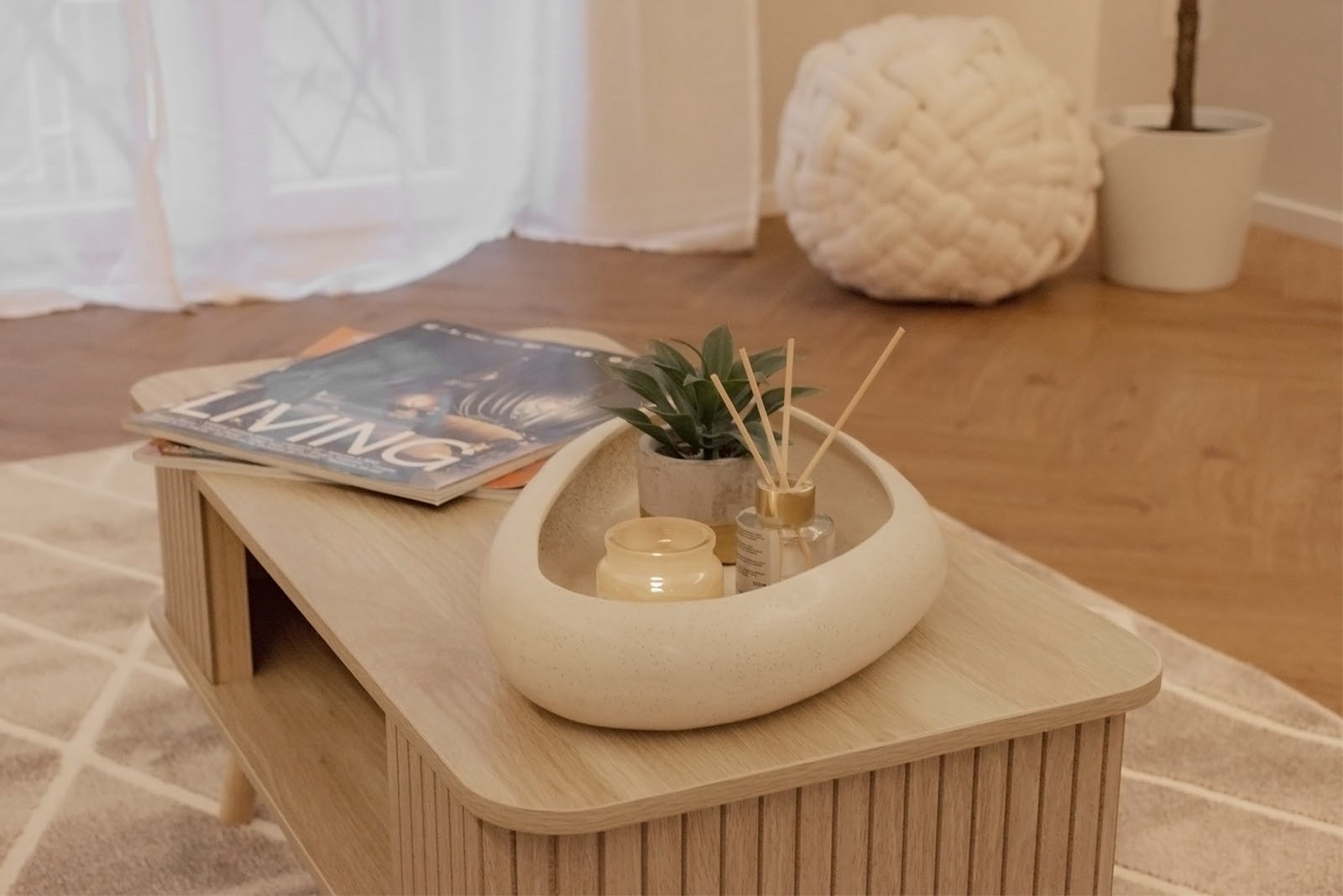6_home-staging-dettaglio-tavolino-soggiorno-moderno-minimalista-design.jpg Dettaglio di tavolino in legno chiaro su tappeto geometrico beige in soggiorno moderno e minimalista; sopra il tavolino vassoio decorativo con pianta verde, candela e diffusore di profumo, accanto a riviste di design, sullo sfondo pouf intrecciato e pianta in vaso vicino alla finestra con tende bianche.