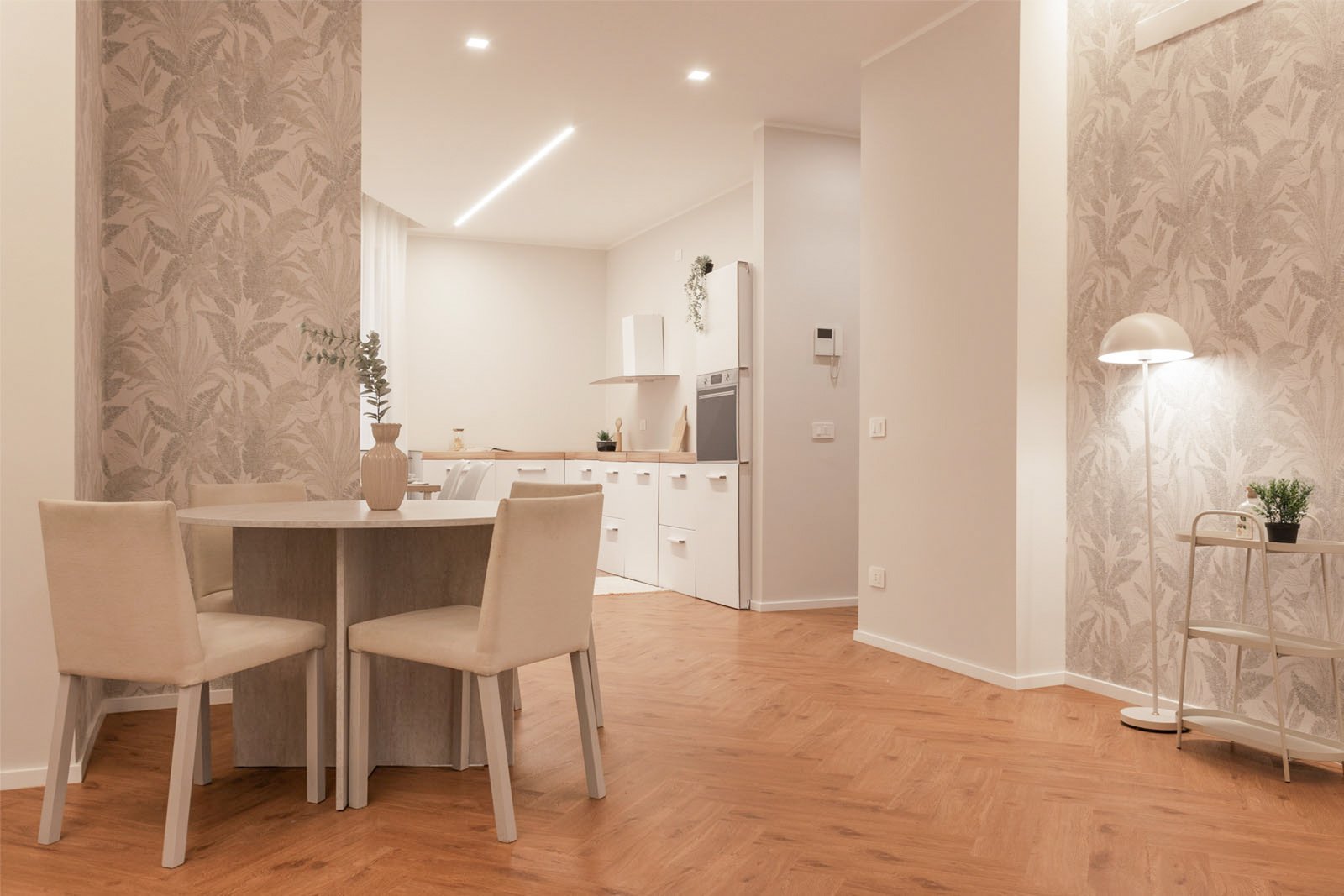 8_home-staging-zona pranzo-cucina-moderno-minimalista-toni-neutri-pavimento-herringbone.jpg Zona pranzo e cucina moderna e minimalista con pavimento in legno a spina di pesce, tavolo rotondo con sedie chiare e vaso decorativo, pareti con carta da parati botanica e illuminazione LED; sullo sfondo cucina bianca in cartone utilizzata per home staging e piccoli accessori; angolo con lampada e pianta verde.