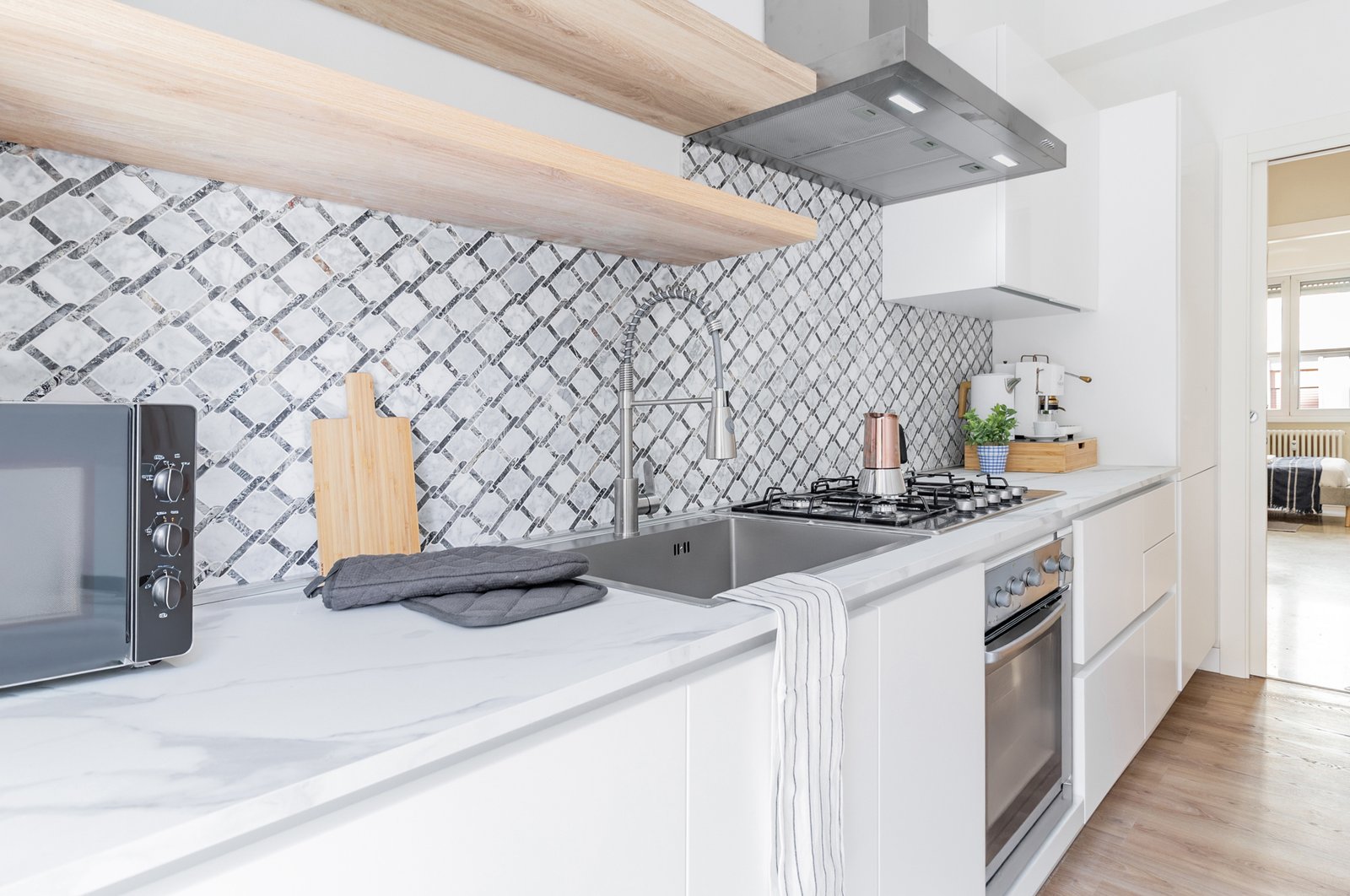 9_cucina-moderna-con-piano-in-marmo-piastrelle-geometriche.jpg Cucina moderna e luminosa con piano in marmo bianco, backsplash a motivi geometrici grigio e bianco, mobili minimalisti, lavello in acciaio e piano cottura con caffettiera.