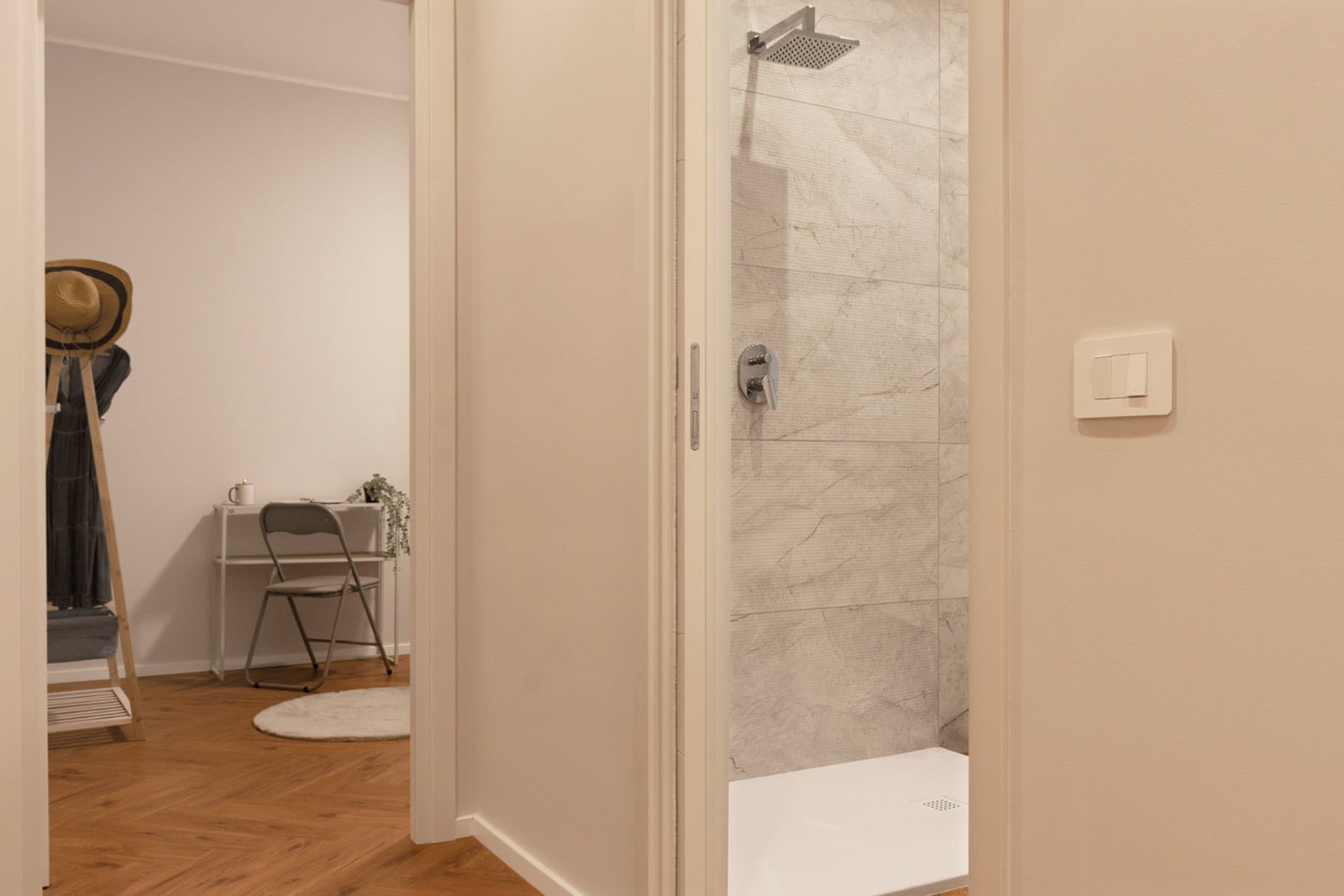 9_home-staging-disimpegno-camera-bagno-moderno-minimalista.jpg Disimpegno moderno e minimalista con pavimento in legno a spina di pesce che collega camera e bagno; sulla destra ingresso alla doccia con rivestimento effetto marmo, sullo sfondo angolo studio con scrivania bianca e sedia pieghevole; ambiente luminoso e rilassante.