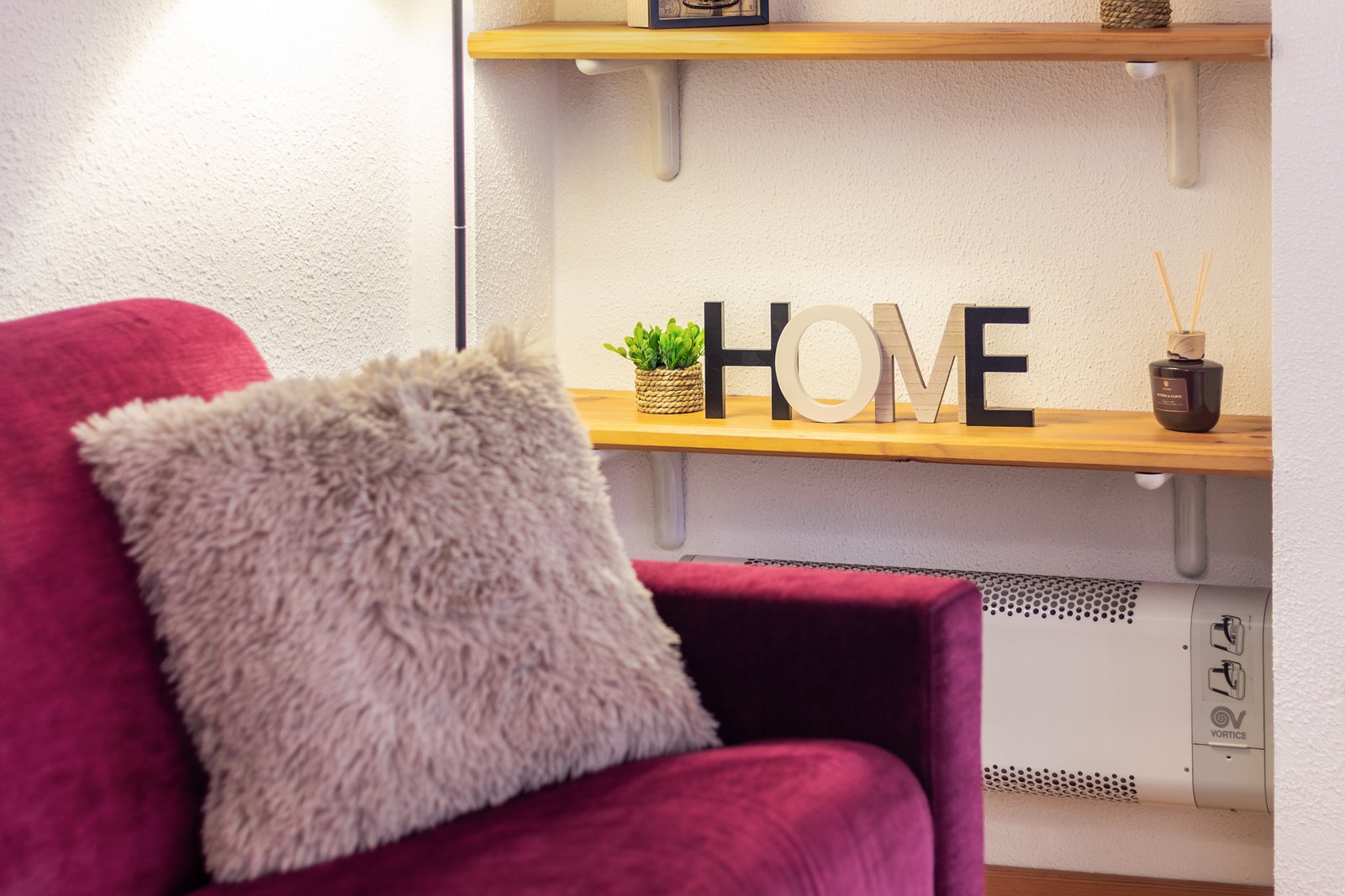 9_relloking-appartamento-montagna-angolo-living-cuscino-home-accogliente-moderno-design.jpg Angolo living accogliente con divano bordeaux e cuscino morbido, mensole in legno con decorazioni e scritta HOME; appartamento moderno e curato nei dettagli.
