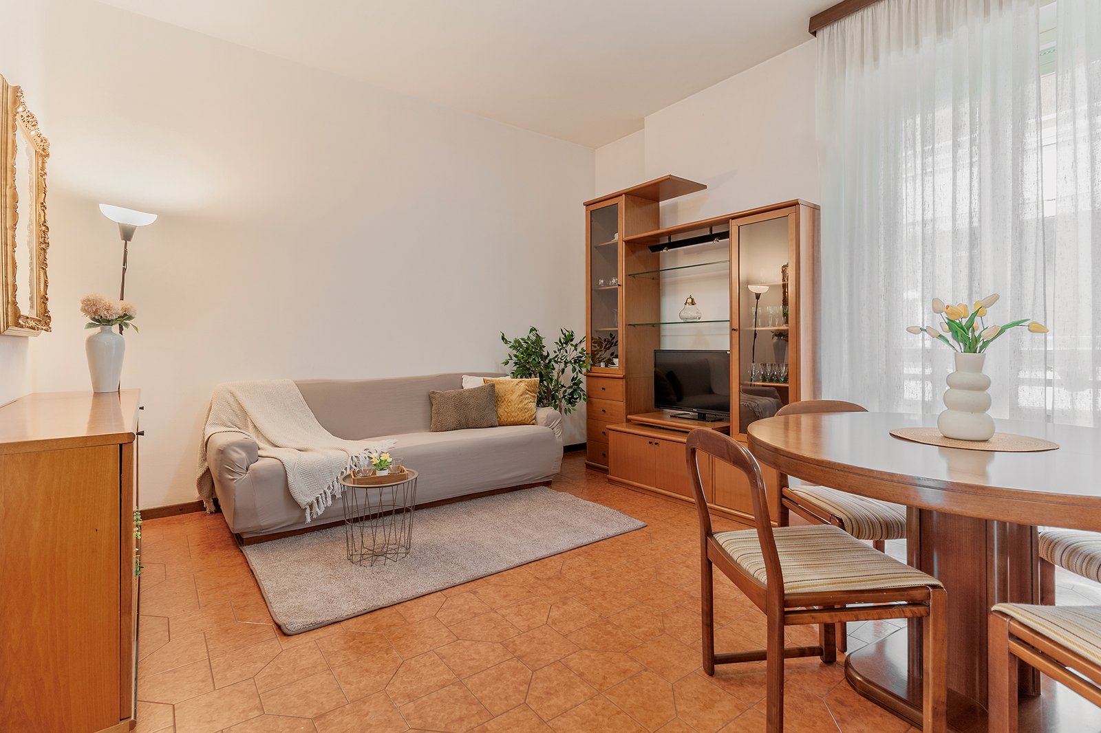 Soggiorno e zona pranzo con arredi in legno e toni caldi, divano grigio chiaro con cuscini dorati e coperta beige, tavolo rotondo con sedie coordinate e vaso di tulipani, mobile TV e credenza in legno, specchio dorato e lampada da terra, pavimento in cotto e tende bianche che diffondono luce naturale.
