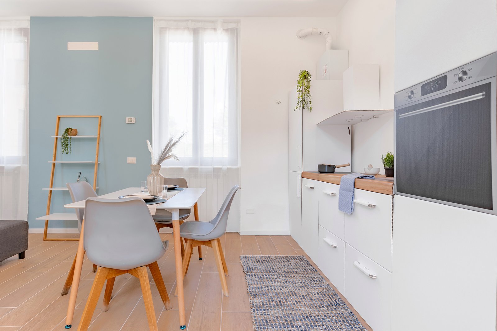 Cucina e zona pranzo moderna e minimalista con pareti bianche e azzurre, pavimento in legno chiaro e arredi dalle linee pulite; tavolo bianco con sedie grigie e gambe in legno, cucina con mobili bianchi e piano in legno, decorazioni con piante verdi e accessori naturali, ambiente luminoso.
